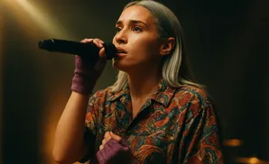 A woman with gray hair sings into a microphone, wearing a colorful shirt and purple fingerless gloves.