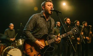 A band performs on stage with a lead singer playing guitar and singing into a microphone, backed by a drummer and saxophonists.