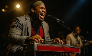 A Black man in a suit and headband plays a lap steel guitar and sings into a microphone on a stage.