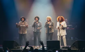 A female vocal group performs on stage under spotlights, with an audience in the foreground.