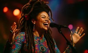 A woman with dreadlocks sings into a microphone, wearing a colorful patterned dress and jewelry.