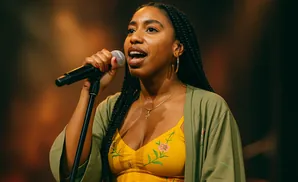 A Black woman with braids sings into a microphone, wearing a yellow floral top and green robe.
