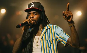 Rapper wearing a baseball cap and colorful striped shirt performs into a microphone, raising his finger in the air.
