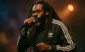 Musician with dreadlocks wearing an Adidas jacket, holding a microphone and performing on stage.