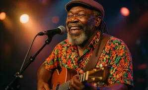 A Black man with dreadlocks and a grey beard, wearing a colorful shirt and cap, laughs while playing an acoustic guitar and singing into a microphone.
