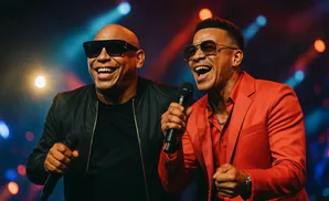 Two men with microphones, one in a black jacket and sunglasses, the other in a red suit, sing on stage with colorful lights.