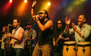 A band performs on stage under warm lights, with a saxophonist, singer, guitarist, and percussionists.