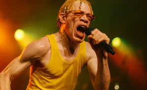 A Black man with albinism, wearing a yellow tank top and sunglasses, sings into a microphone on stage with stage lighting.