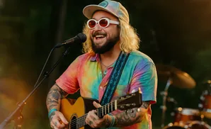 A man with blonde hair, a tie-dye shirt, and round white sunglasses plays an acoustic guitar and sings into a microphone.