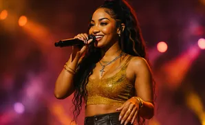 A woman with long, dark, wavy hair, wearing a gold sequined top and black pants, sings into a microphone on stage.