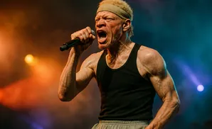 Singer with albinism, wearing a black tank top and headband, performs with a microphone on a smoky stage.