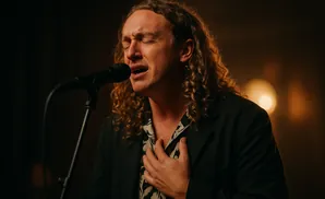A male singer with curly hair sings passionately into a microphone, his eyes closed and hand on his chest.