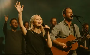 Woman singing with a raised hand, man playing guitar, and others singing in the background.