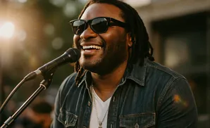 A man with dreadlocks and sunglasses smiles into a microphone, wearing a denim shirt.