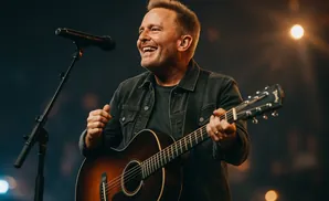 A man with short blonde hair plays an acoustic guitar and sings into a microphone on a dimly lit stage.