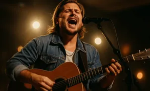 A man sings into a microphone while playing an acoustic guitar under warm stage lights.