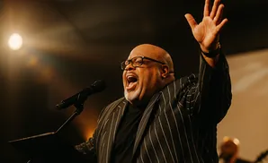 A Black man in a pinstripe suit sings passionately into a microphone, with one arm raised.