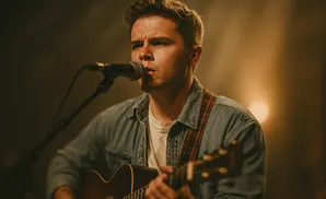 Man in denim shirt playing acoustic guitar and singing into a microphone.