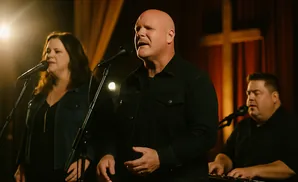 A bald man sings into a microphone, with a woman and a man playing piano beside him.