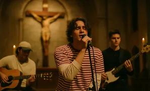 A band performs in a church, with a crucifix in the background. The lead singer, in a striped shirt, sings into a microphone.
