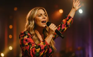 Woman singing into a microphone on stage, wearing a floral dress and raising her arm.