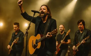 Lead singer of a band performs, raising a fist in the air while playing an acoustic guitar.