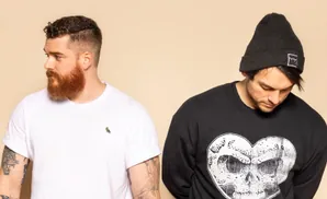 Two men against a beige background. The man on the left wears a white t-shirt and has a beard. The man on the right wears a black beanie and sweatshirt …