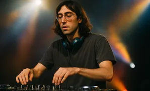 A DJ with long brown hair and glasses, wearing headphones, mixes music on a DJ setup in a dimly lit room with colorful stage lights.