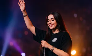 A woman sings into a microphone with her arm raised in a concert with colorful lights.