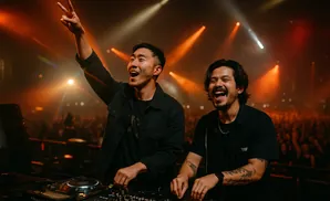 Two DJs in black shirts joyfully perform for a large crowd under vibrant stage lights.