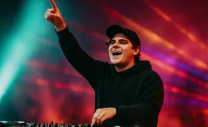 A DJ in a black hoodie and cap points upwards while operating a DJ set with a colorful light show in the background.