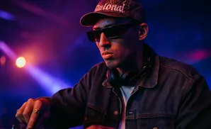 A DJ wearing a baseball cap and sunglasses, with headphones around his neck, plays a mixing board in a club with purple and orange lighting.