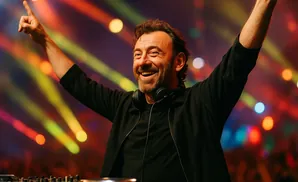 DJ with arms raised, smiling, at a colorful, light-filled concert.