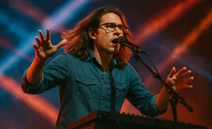 Man with long hair and glasses singing into a microphone on stage, with blue and orange stage lights.