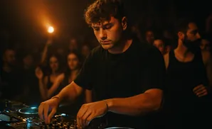 A DJ smiles while working a console at a concert, illuminated by a bright spotlight.