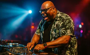 A Black DJ in a tropical shirt smiles while operating turntables under blue and red stage lights.
