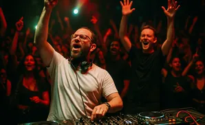 A DJ with his arm raised in celebration at a lively concert, with a cheering crowd in the background.