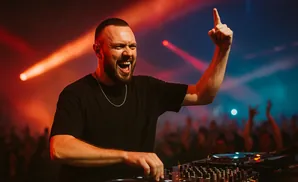 DJ in black t-shirt with a gold chain, enthusiastically performing at a console with red and blue lights.