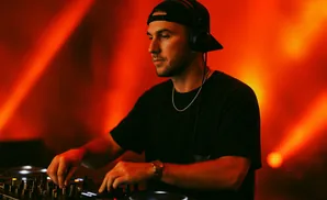 DJ wearing headphones and a black cap mixing music on a turntable with red stage lighting.