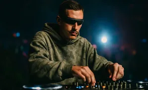 A DJ wearing futuristic glasses and a green hoodie plays music on a console in a dark room.