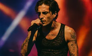 A male singer with tattoos performs on stage, holding a microphone and wearing a black tank top.