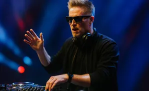 A DJ in sunglasses and headphones plays music on turntables, with hands reaching out to the equipment.