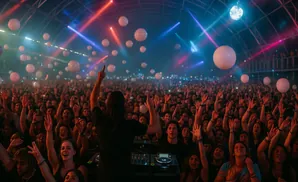 A DJ with arms raised plays to a large, cheering crowd at a concert, with lights and balloons overhead.