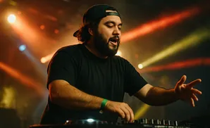 A DJ in a black t-shirt and baseball cap gestures with his hands while mixing music on a Pioneer DJ setup.