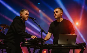 Two men perform music on stage with keyboards and a laptop, illuminated by blue and orange stage lights.