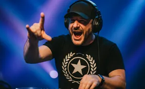 DJ in black cap and headphones, excitedly gestures with hand raised, playing music.