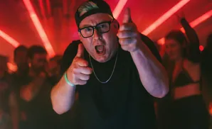 Man in a black shirt and baseball cap points at the camera with a green bracelet on his arm.