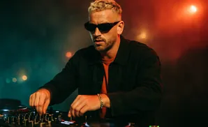 DJ with blonde hair, sunglasses, and a black jacket, adjusting controls on a DJ mixer in a dimly lit club with red and green lighting.