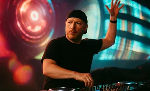 DJ in a black beanie and t-shirt, hands on DJ equipment, with a blurred, colorful background.
