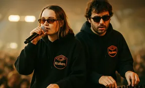 Two DJs wearing black hoodies with an orange logo perform on stage, with one singing into a microphone.